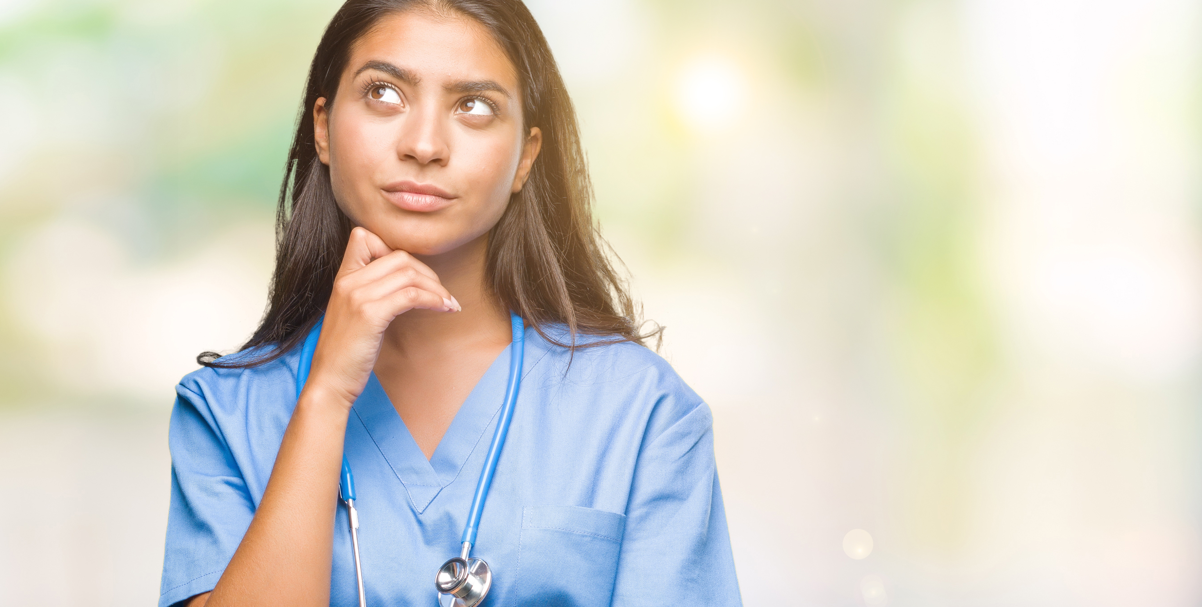 A nurse in blue scrubs with a stethoscope resting a hand on her chin, looking upward in thought.